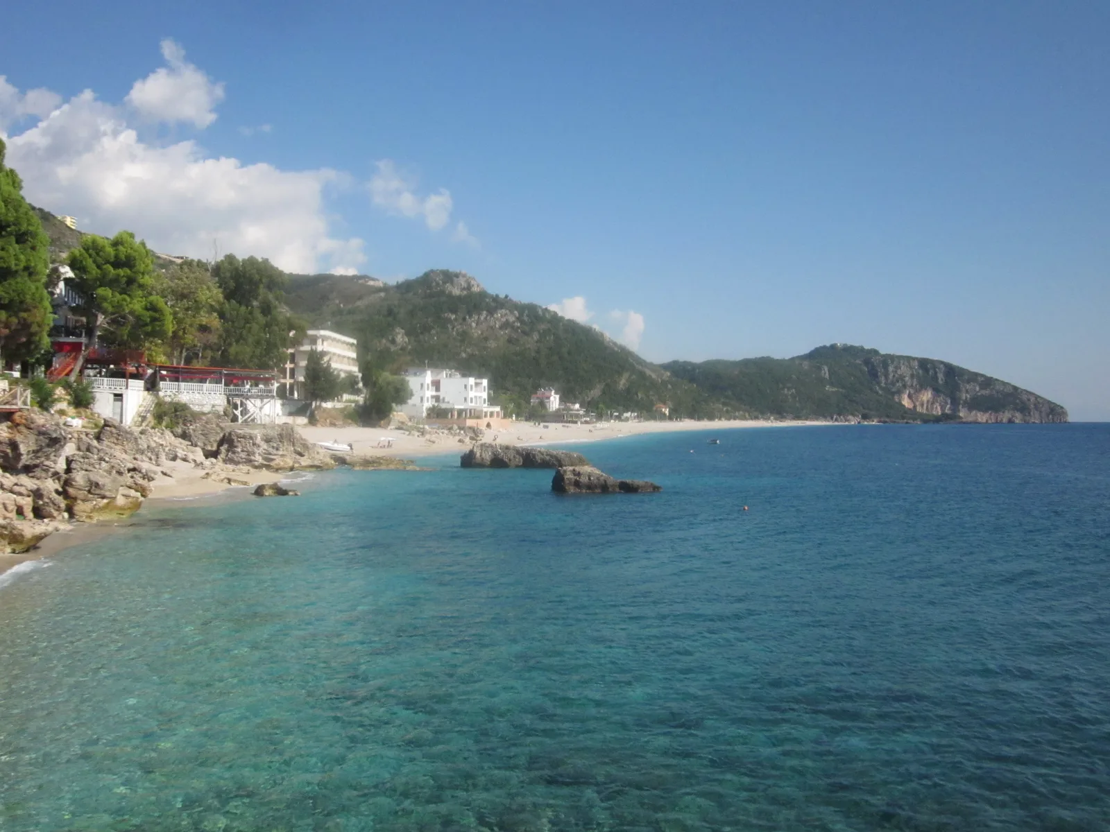 The pale pebble shore at Dhërmi curving away below wooded slopes, clear turquoise shallows over rock and a headland closing the bay
