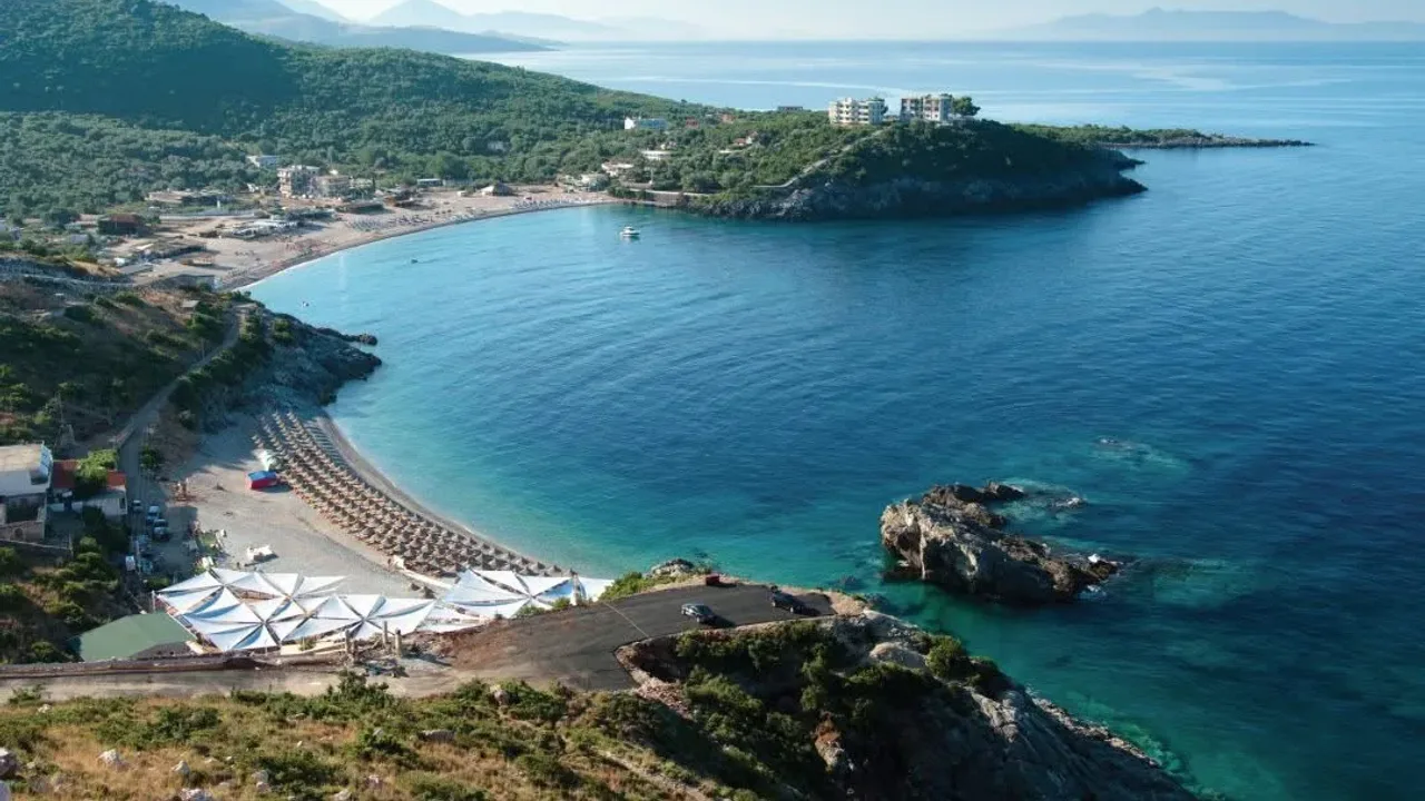 Aerial view of Jale Beach, a curved cove with beach clubs and a rocky headland on the Albanian Riviera