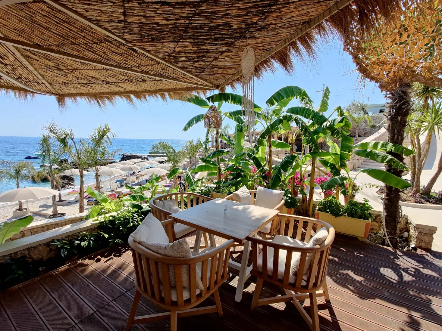 A reed-shaded terrace of wicker chairs above straw parasols and turquoise water at Flow Beach on the Dhërmi shore