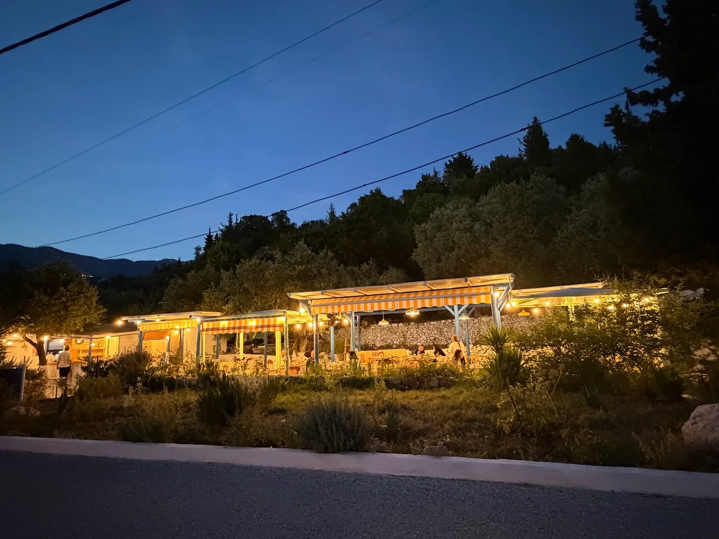 The yellow-striped awnings and string lights of The Jester's Taverna lit up at dusk beside the Himarë–Livadh road