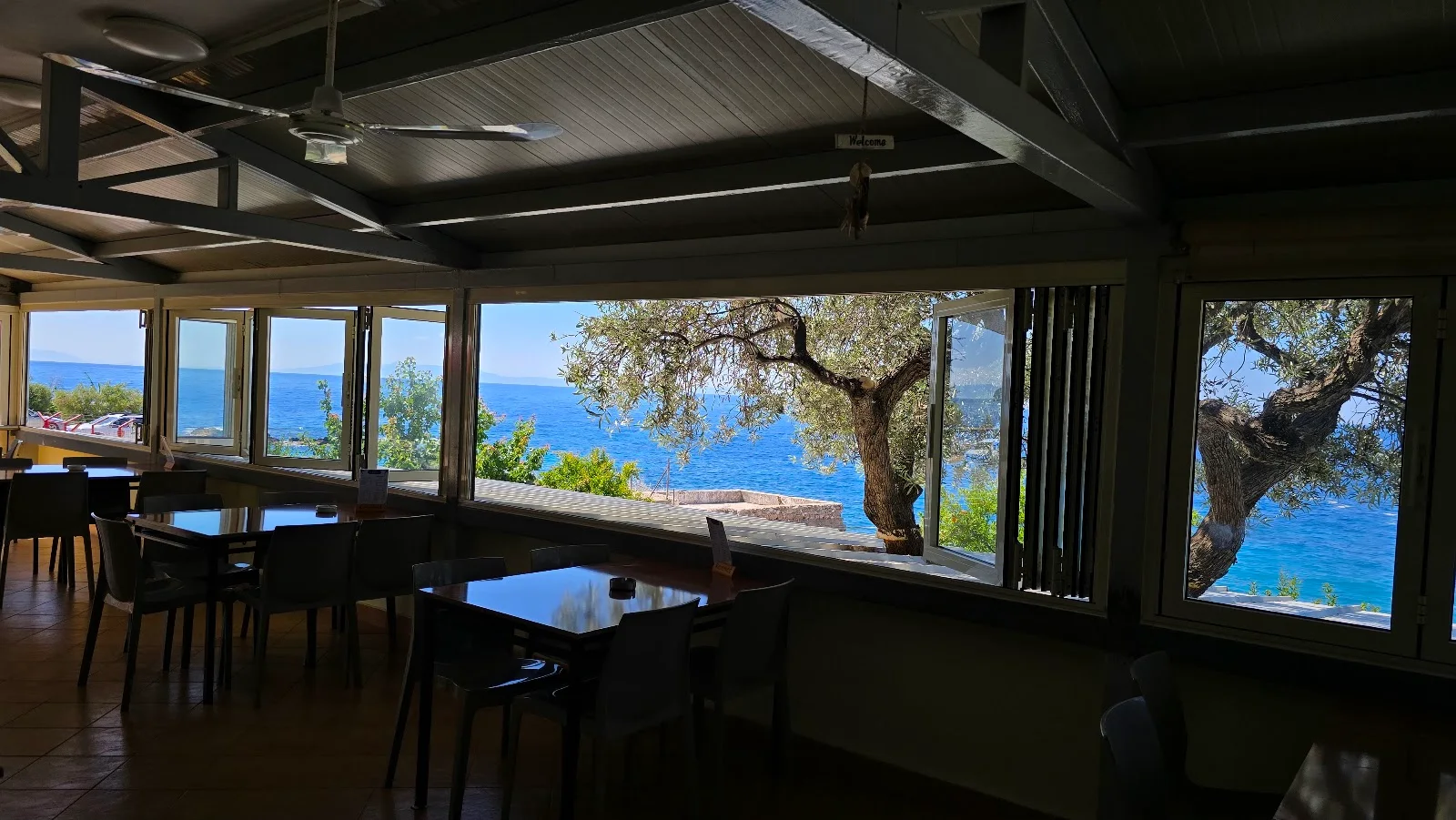 The glazed sea-view dining room of Ristorante Pano Marko with an olive tree framing the Ionian at Dhërmi