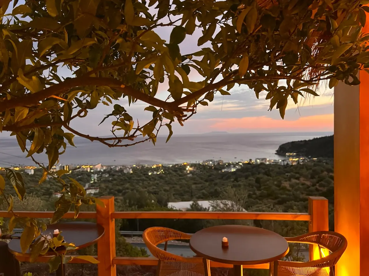 Sunset over the bay framed by a tree, with bistro tables on the hilltop terrace at Mirage cocktail bar above Livadhi