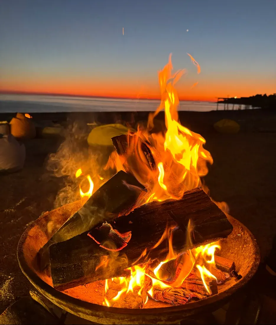 A beach fire pit blazing at dusk with beanbag loungers on the sand at Salinas Beach Bar in Dhërmi