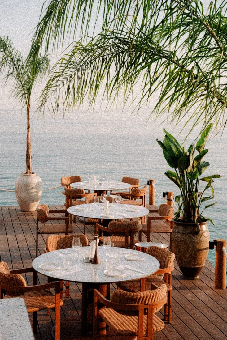 Round terrazzo tables and rope-and-teak armchairs set for dinner on the sea-edge timber deck at Cavos Mediterranean Experience, Dhërmi