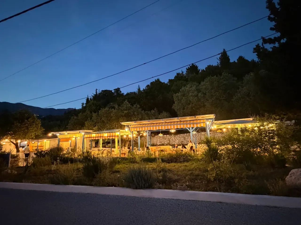 The yellow-striped awnings and string lights of The Jester's Taverna lit up at dusk beside the Himarë–Livadh road