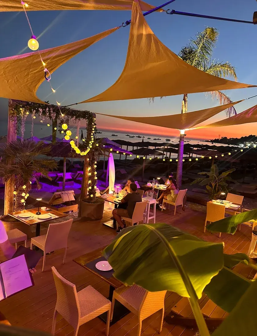 The sail-shaded beach terrace of olio lit with string lights at sunset over the Dhërmi shore