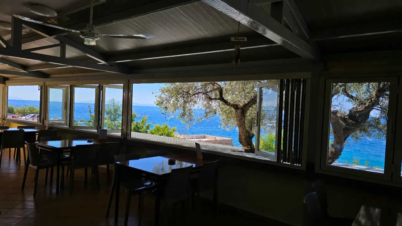 The glazed sea-view dining room of Ristorante Pano Marko with an olive tree framing the Ionian at Dhërmi