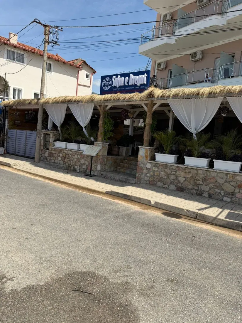 The thatched, white-curtained street terrace and sign of Sofra e Bregut traditional grill house in Himarë