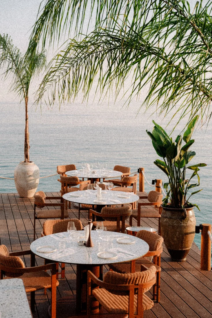Round terrazzo tables and rope-and-teak armchairs set for dinner on the sea-edge timber deck at Cavos Mediterranean Experience, Dhërmi